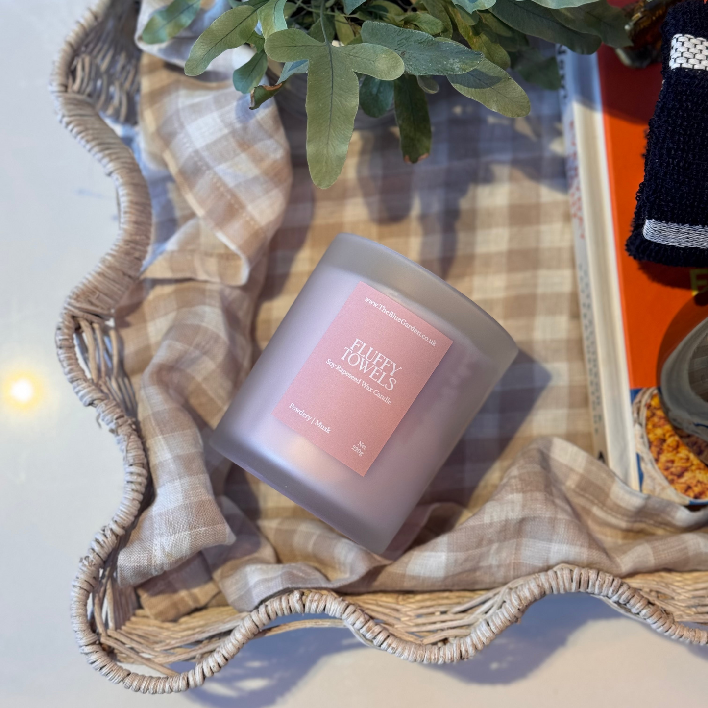 Candle with a pink label on a woven surface with greenery and a plaid cloth.