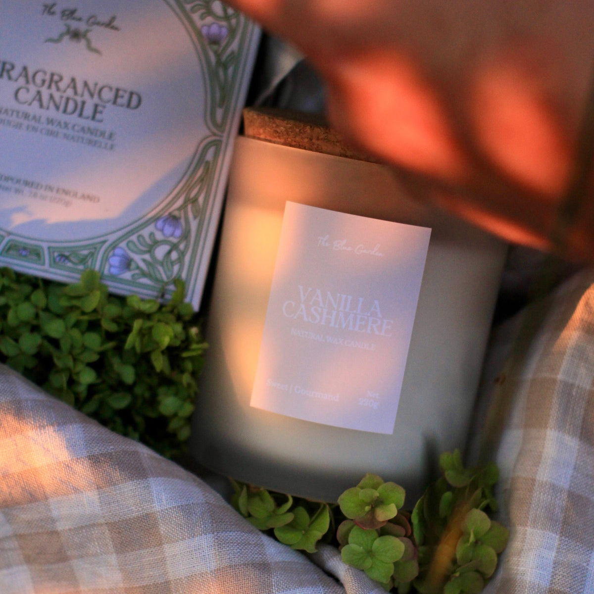A hand holds a woven basket over a lit scented candle in a gift basket lined with a beige plaid blanket, surrounded by green leaves. The candle label reads 'Vanilla Cashmere' and the box says 'Fragranced Candle' by The Blue Garden