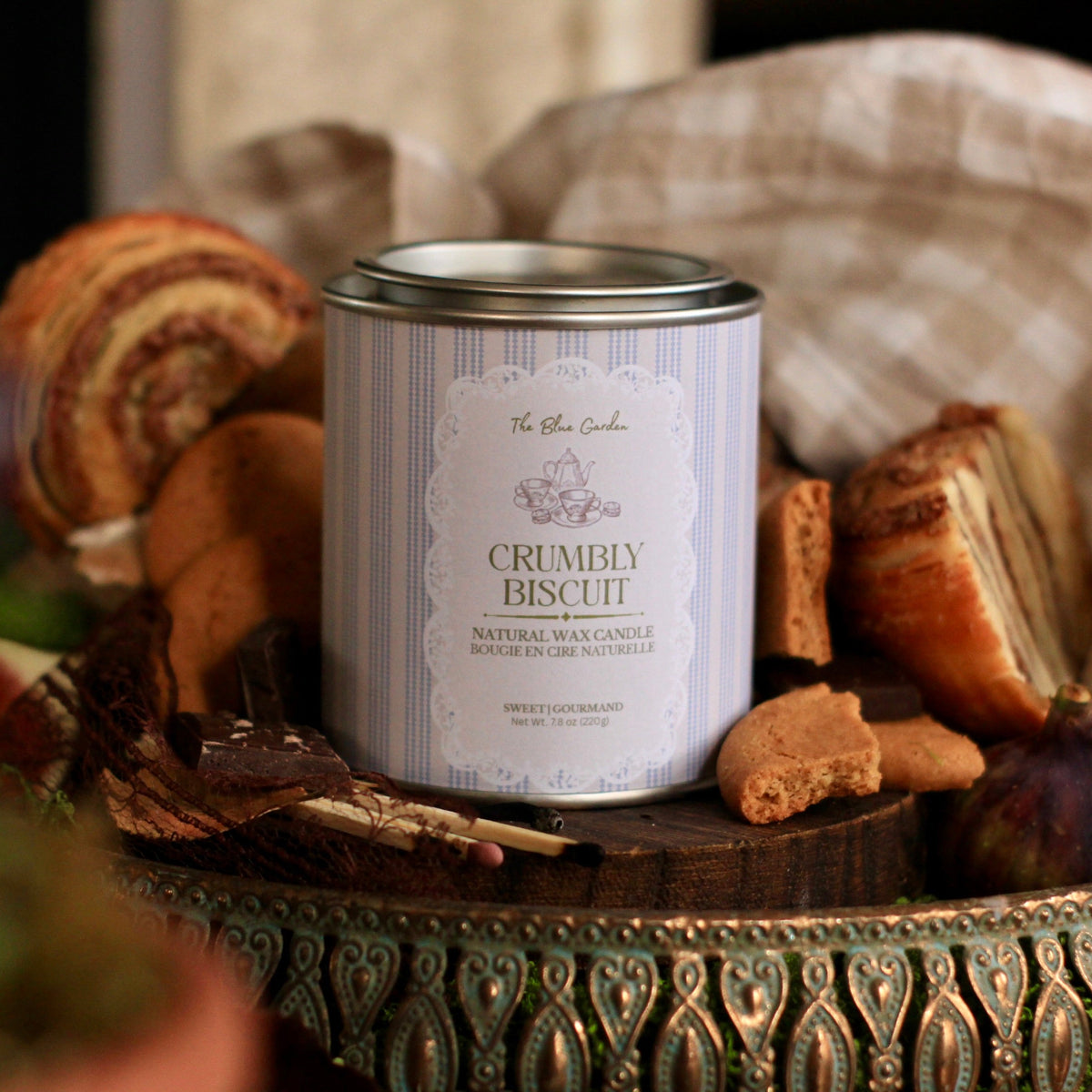 A tin candle labeled 'Crumbly Biscuit' by The Blue Garden, styled on a decorative tray with cookies, bread, and a checkered cloth in the background.