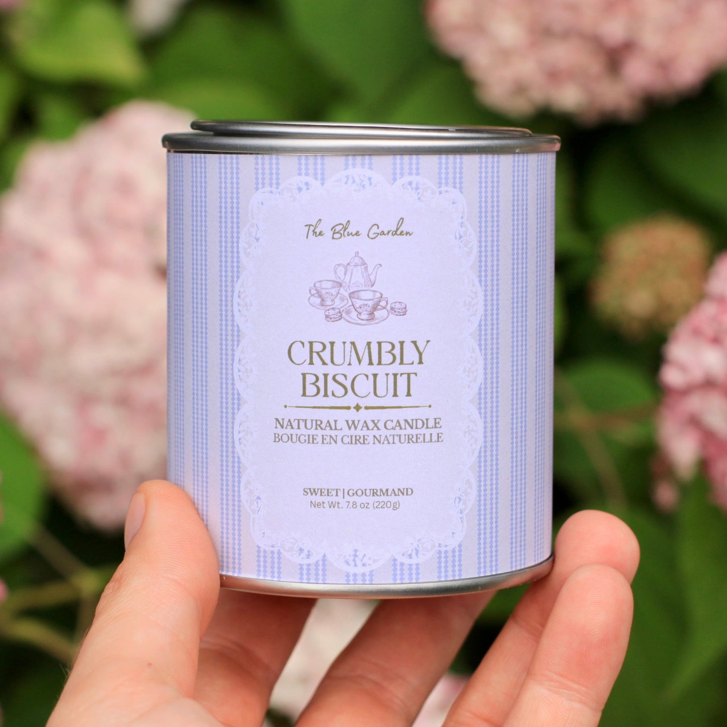 Hand holding a candle can labeled 'Crumble Biscuit' against a floral background