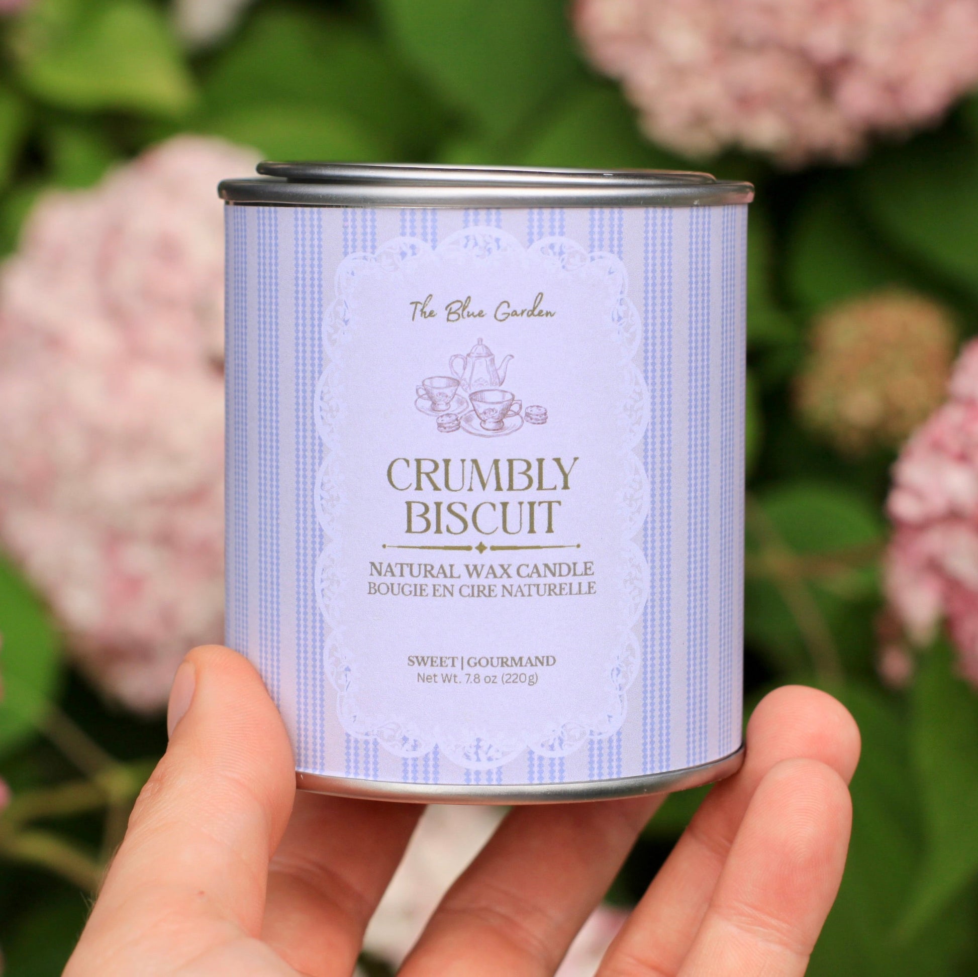 Hand holding a candle can labeled 'Crumble Biscuit' against a floral background
