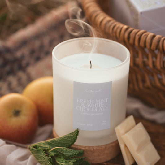 A blue scented candle in a clear glass jar sits on a wooden cork lid on a rustic table, surrounded by mint leaves, white chocolate pieces, and red apples. A wicker basket with a gift box is blurred in the background.