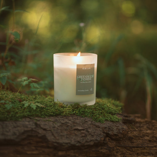 A lit white candle in a clear glass jar sits on moss atop a log in an outdoor forest setting with a soft, blurred green background.
