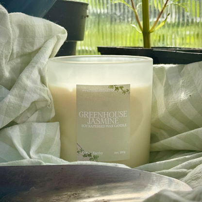 scented soy candle labeled 'Greenhouse Jasmine' on a wooden surface with plants in the background and greenhouse tools