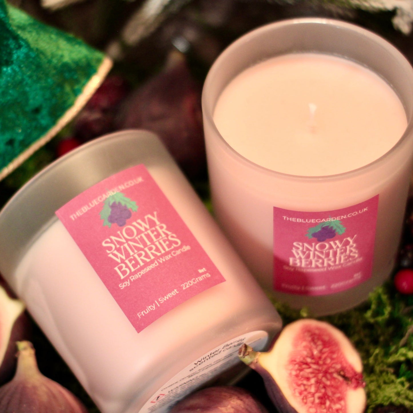 Two candles labeled 'Snowy Winter Berries' surrounded by figs and berries on a textured mossy tray