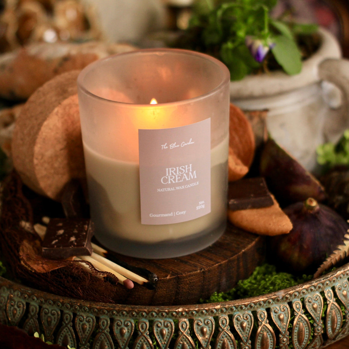 A lit candle in a frosted glass jar sits on a decorative tray surrounded by figs, chocolate pieces, moss, and rustic props. The candle label reads 'The Blue Garden' and 'Irish Cream'.