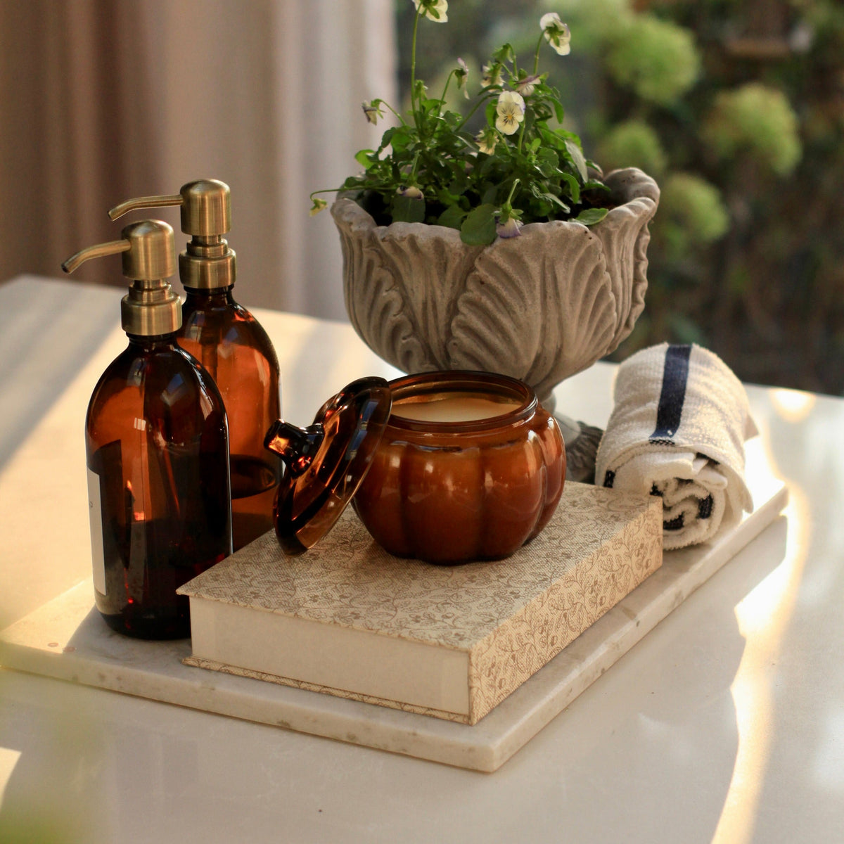 A styled bathroom or spa setup on a white tray, featuring amber glass pump bottles, a small amber jar, a textured stone planter with greenery, and a rolled towel, set against a softly blurred window background.