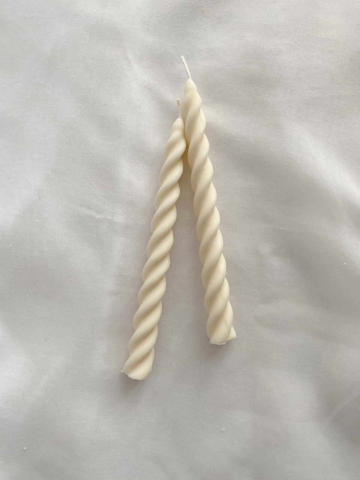 Two cream-colored twisted spiral taper candles standing upright on a white fabric background.