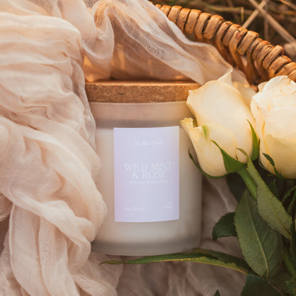 A jar candle with a natural cork lid and a white label reading 'Wild mint & rose' is styled in a woven basket with cream fabric and white roses.