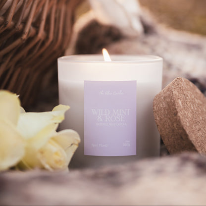 A lit white candle with a lavender label reading 'Wild Mint & Rose' sits on a blanket outdoors, styled with a wicker basket, white flower petals, and a cork lid.