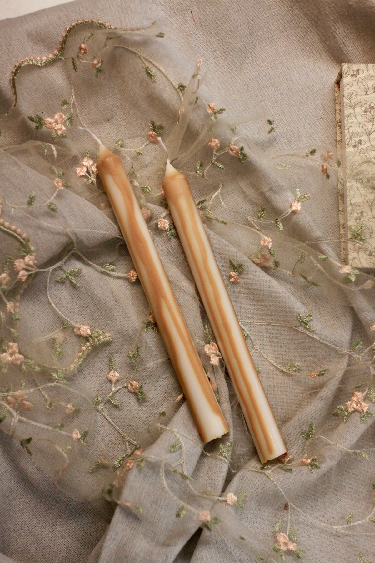Two large size tapered caramel drip candles with a warm tan and cream marbled pattern, resting on a dusty gray-beige embroidered sheer fabric.