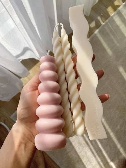 A hand holds three sculptural wax candles in a home setting with sunlight and curtains in the background. The candles include a pink ribbed bar candle, a cream twisted taper candle, and a cream wavy taper candle.