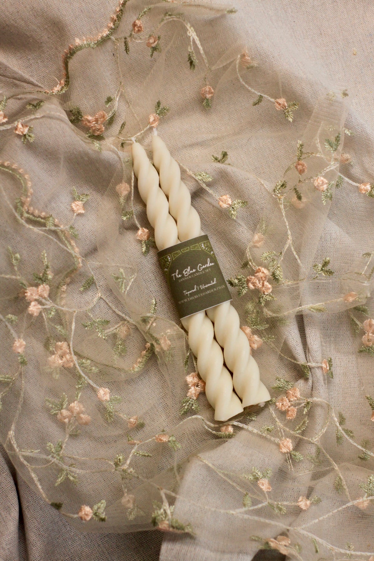 Two twisted taper candles wrapped in a dark olive green paper band, placed on draped beige fabric with delicate floral embroidery.